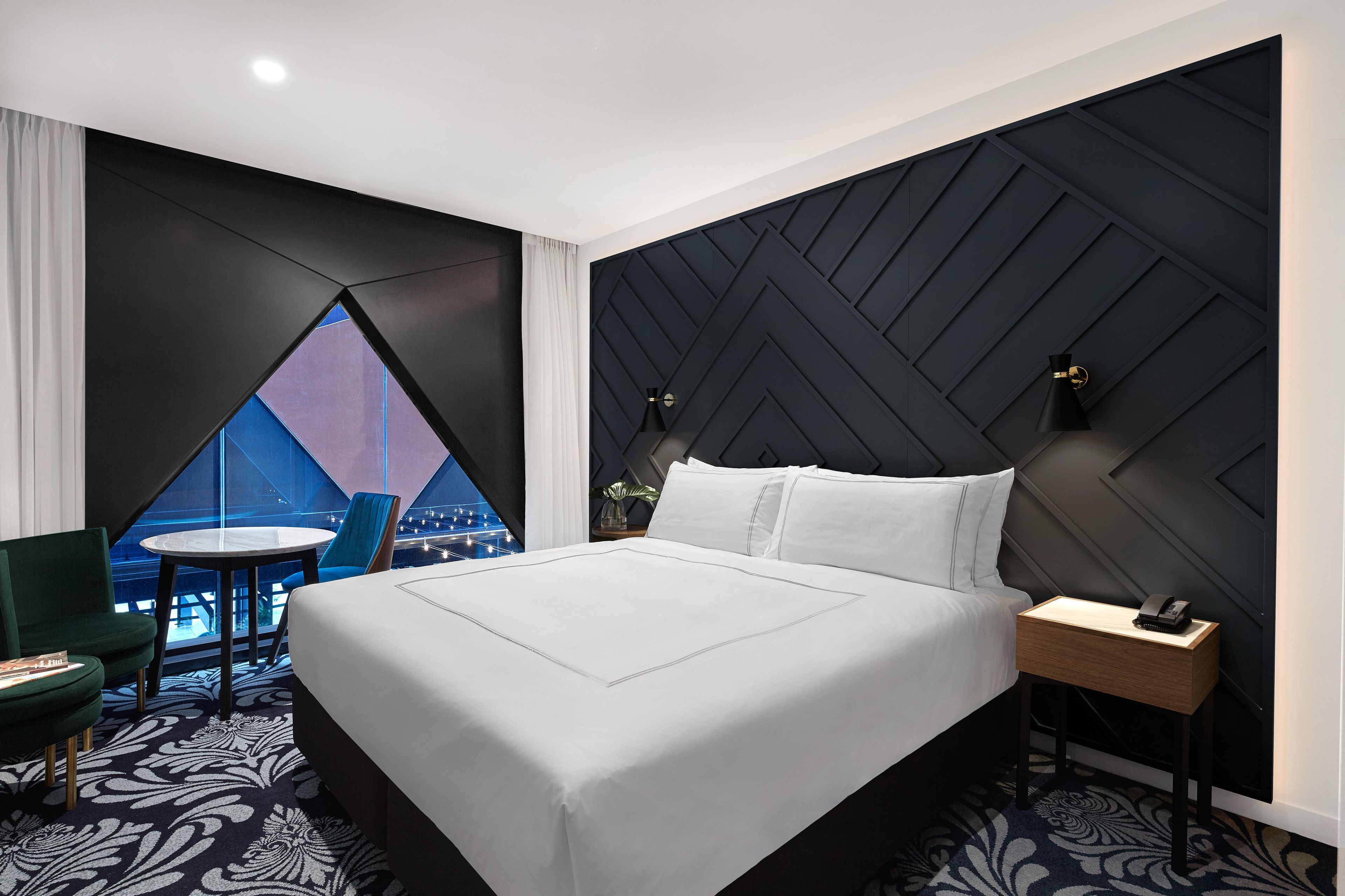 A modern hotel room with a large bed, a view of the cityscape, a small round table, and a chair.