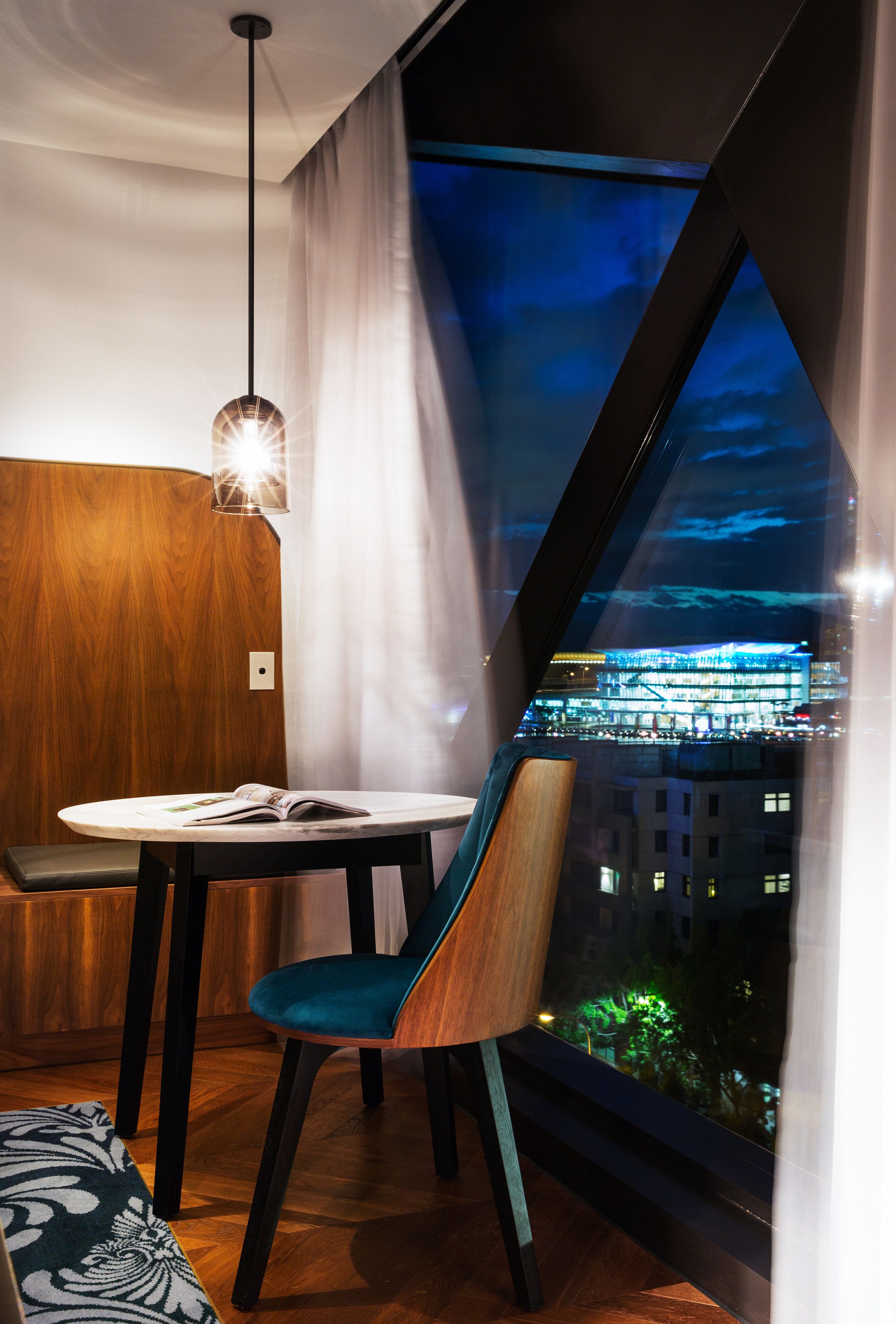 A modern room with a large window, a round table, a blue chair, and a hanging light fixture.