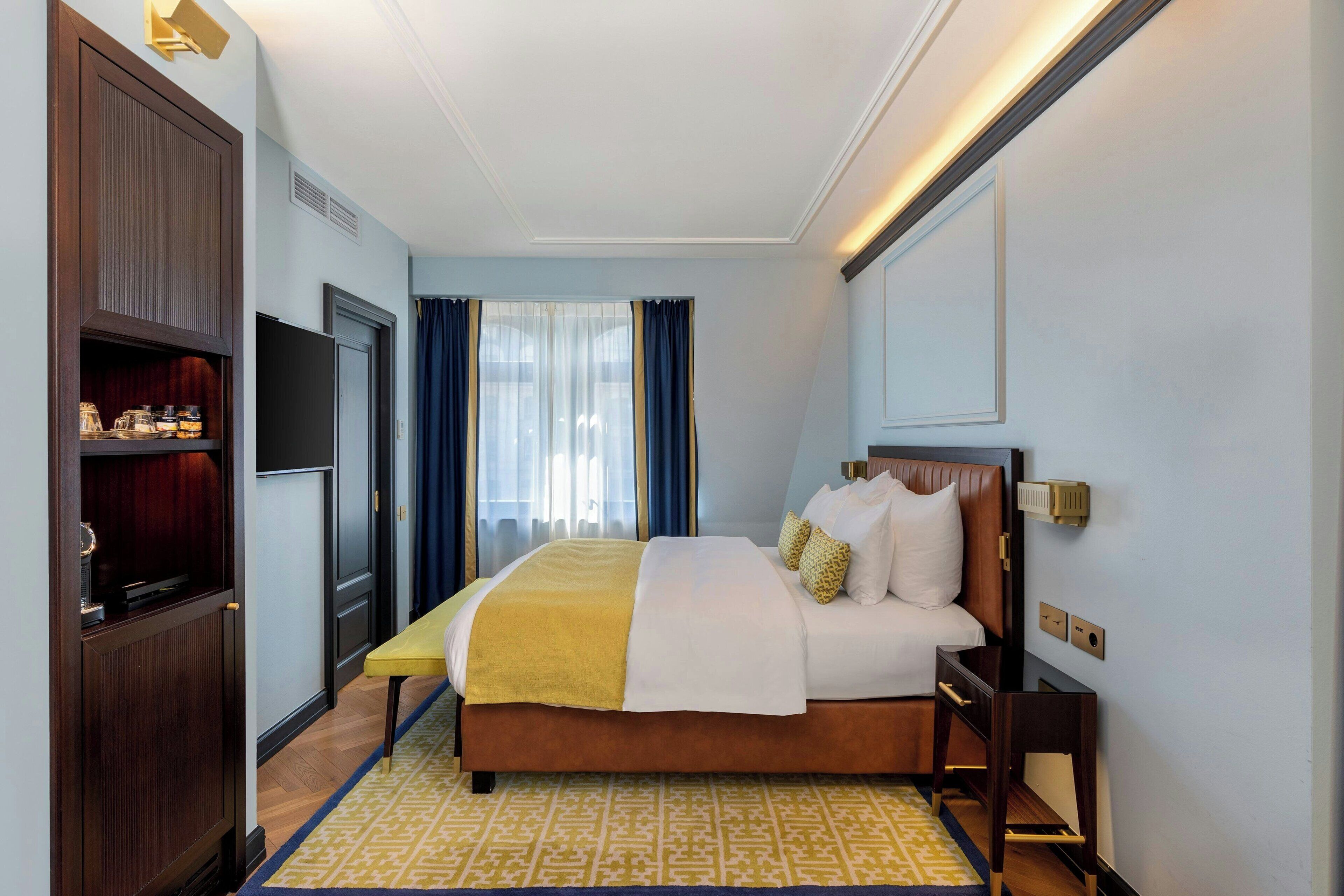 Une chambre d’hôtel avec un grand lit, une table de chevet, une armoire, une télévision et une fenêtre avec des rideaux.