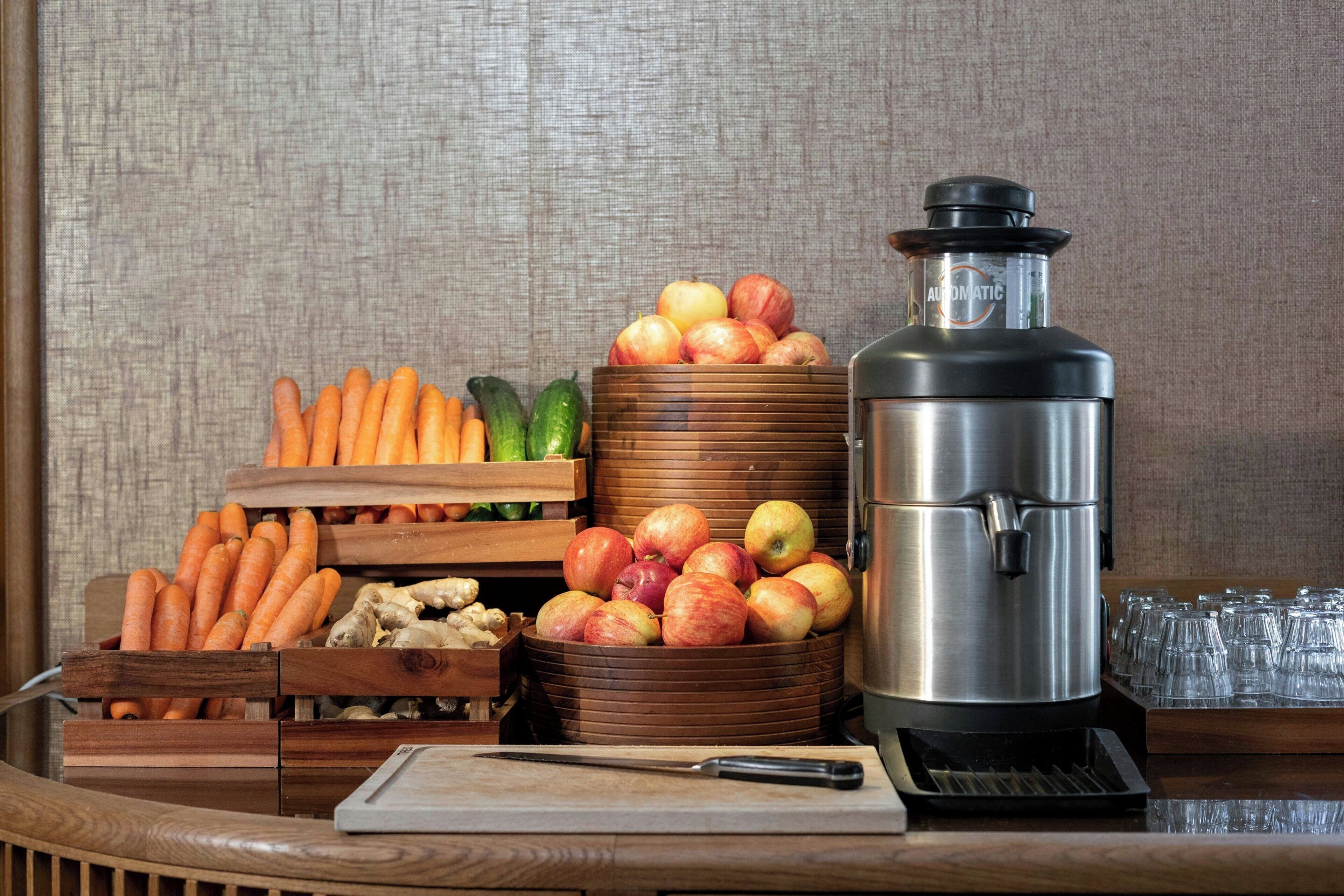 Una mesa de madera con una tabla de cortar, un cuchillo, una cesta de frutas con manzanas y zanahorias, un pepino y un electrodoméstico de acero inoxidable.