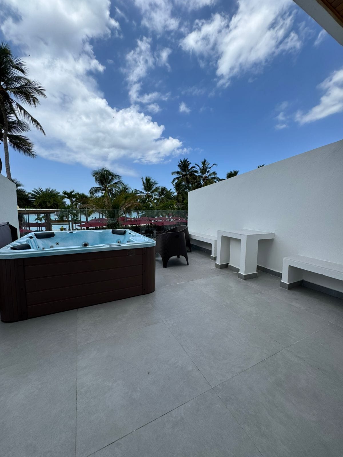 Una terraza en la azotea con jacuzzi, zonas de estar y vistas a palmeras bajo un cielo parcialmente nublado.