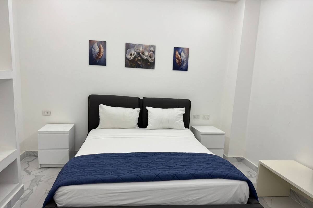 A modern bedroom with a large bed, white bedding, and a blue quilt. There are two bedside tables with lamps, and three wall-mounted paintings above the bed.