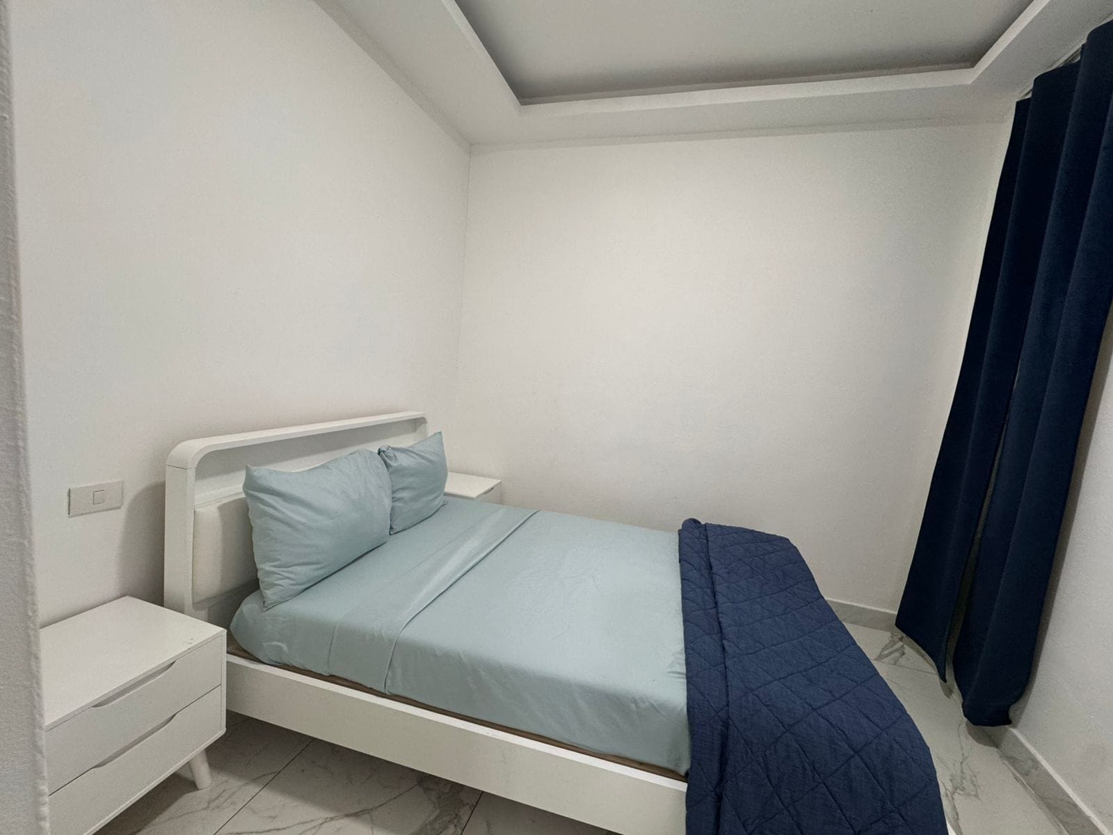 Una habitación pequeña con cama individual, cabecera blanca, mesita de noche y cortina azul oscuro.