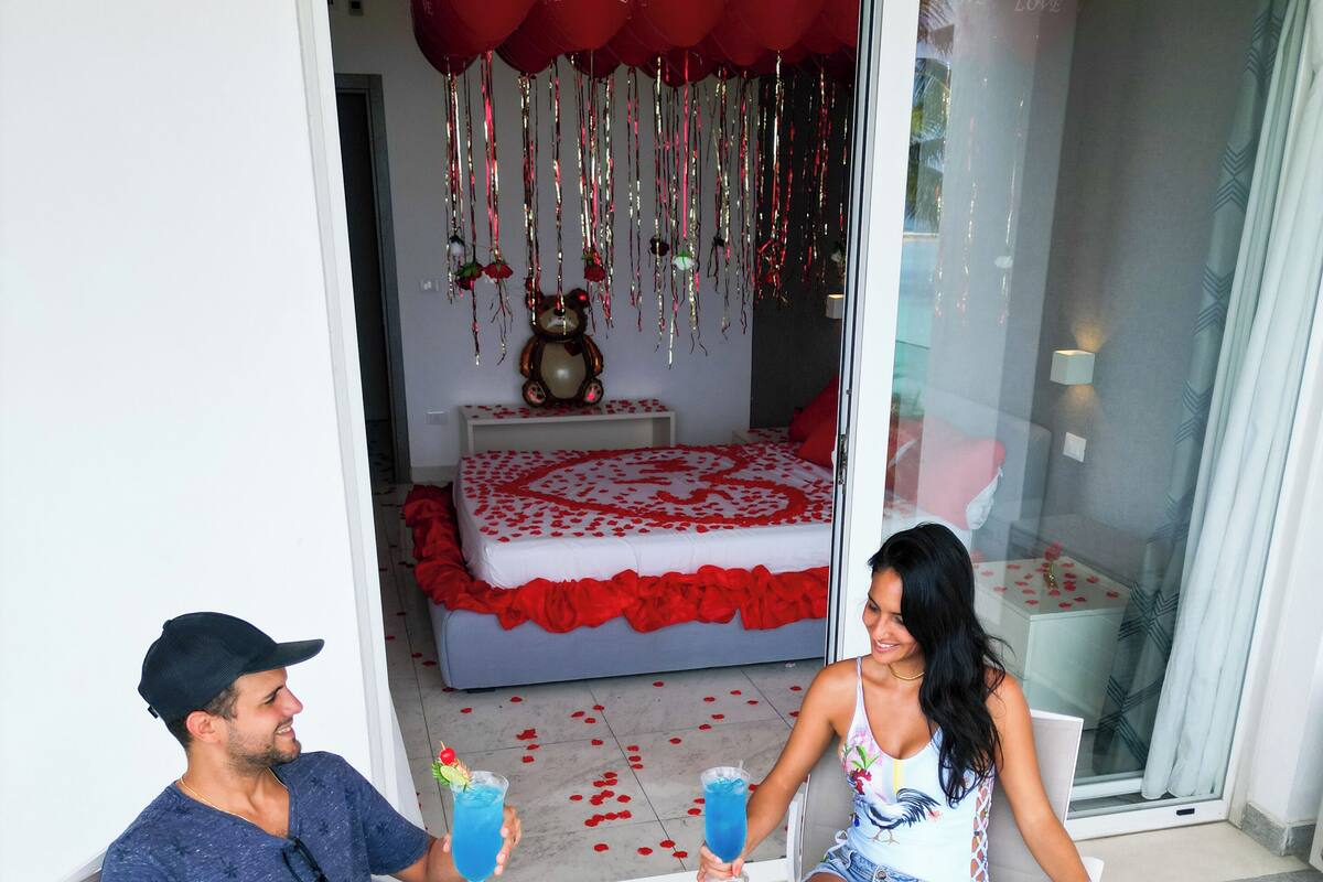 A couple seated at a table with drinks, a bed with red and white bedding, and a teddy bear on the nightstand.