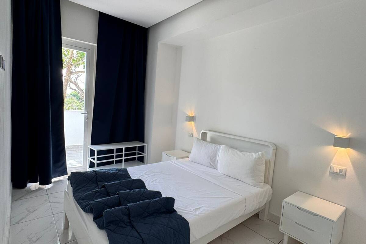A modern bedroom with a large bed, white walls, and a view of the outdoors through a window with dark curtains.