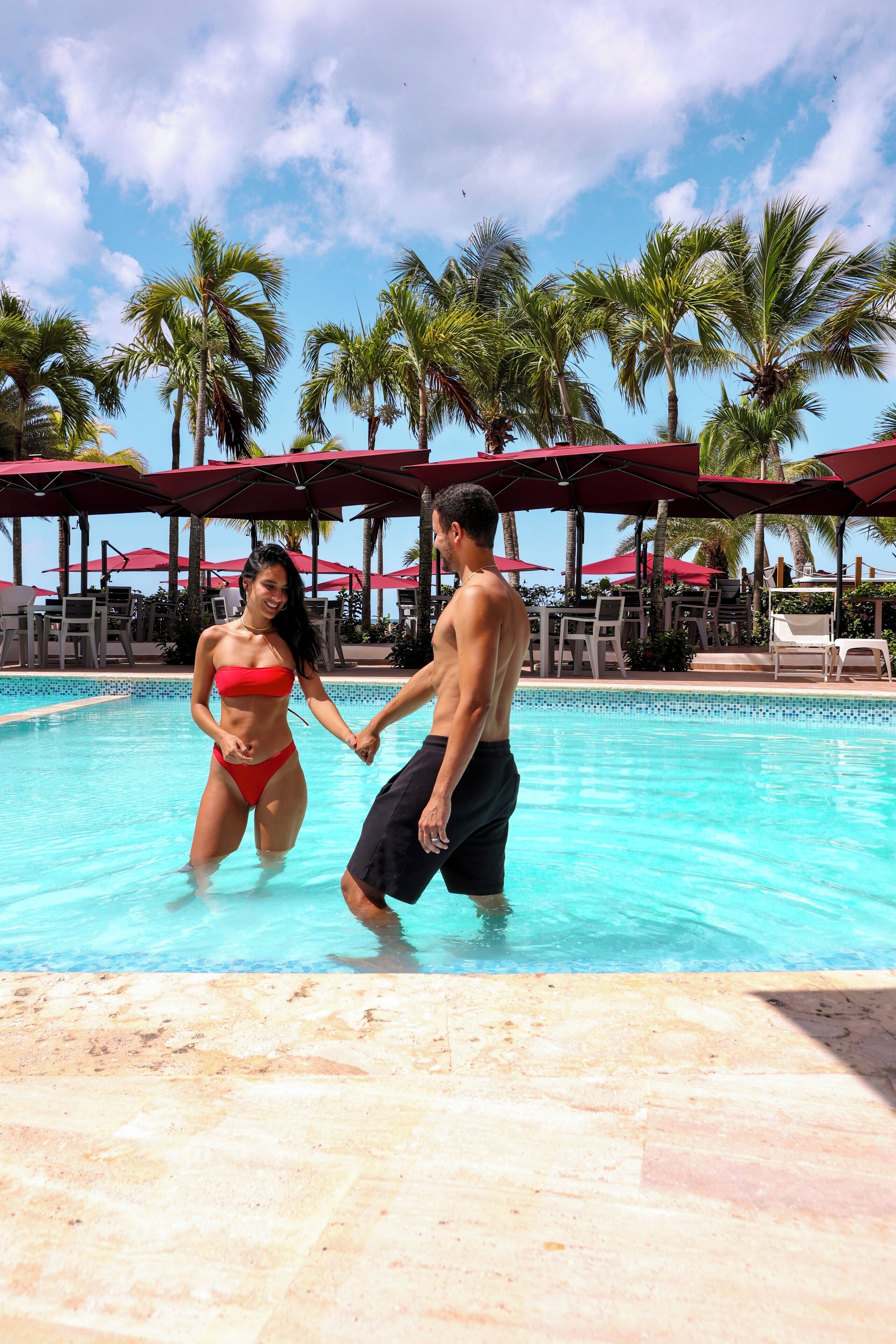 A couple holding hands in a pool with palm trees and umbrellas in the background.