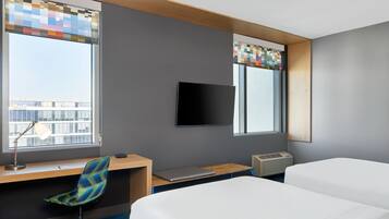 A hotel room with a desk, two beds, a TV, and a window with a view of buildings.
