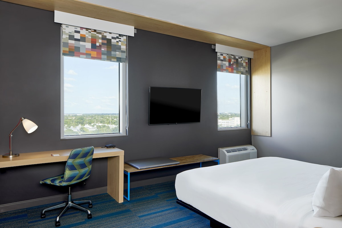 Une chambre d’hôtel équipée d’un lit, d’un bureau, d’une chaise, d’une télévision et de fenêtres avec des stores.
