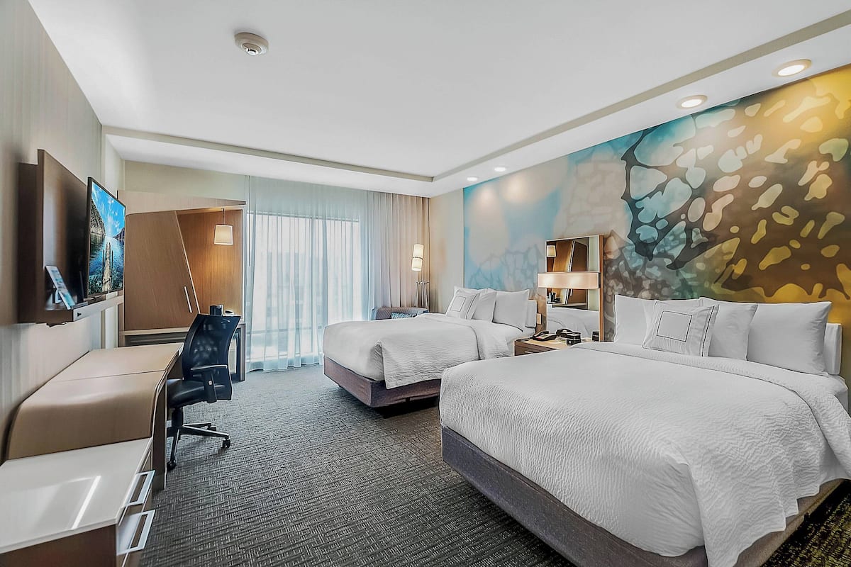Ein Hotelzimmer mit zwei Betten, einem Schreibtisch, einem Stuhl, einem Fernseher und einem großen Fenster mit Vorhängen.
