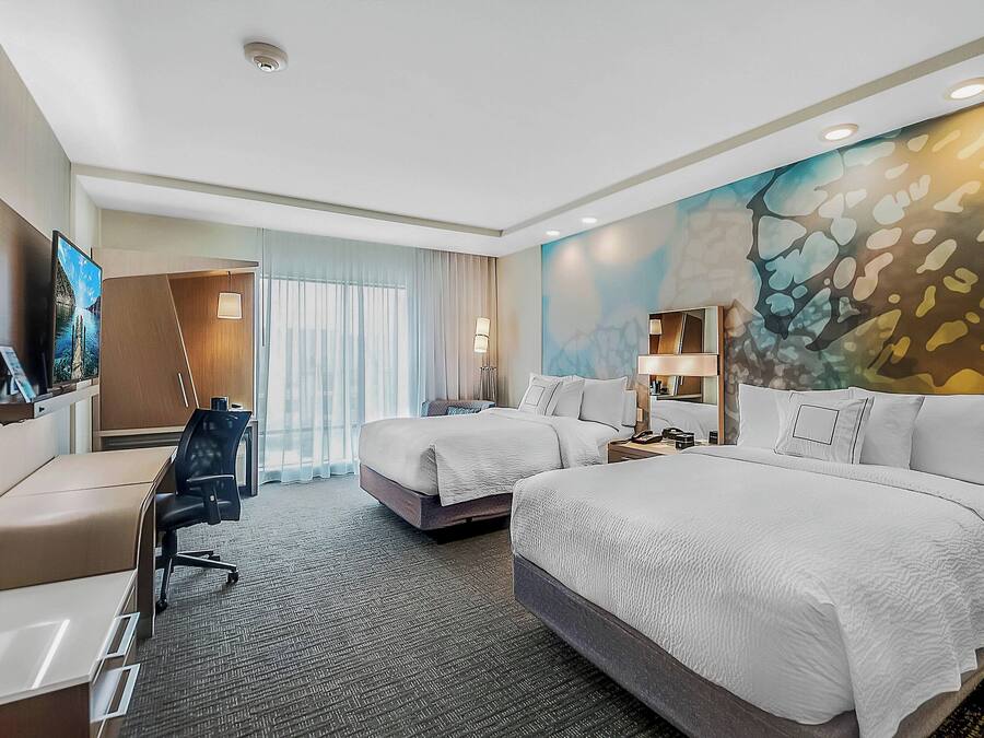 A hotel room with two beds, a desk, a chair, a TV, and a large window with curtains.