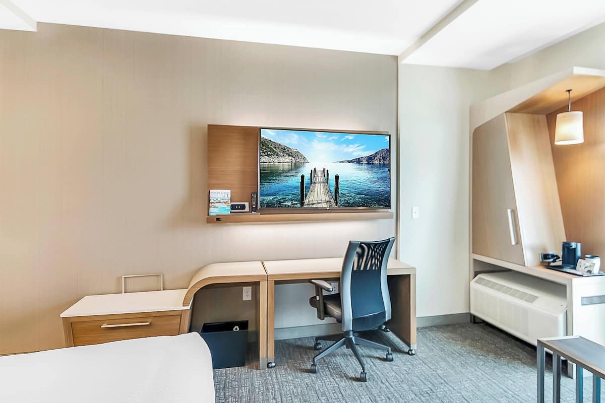 Ein Hotelzimmer mit einem Bett, einem Schreibtisch, einem Stuhl, einem an der Wand befestigten Fernseher und einem Fenster mit Blick auf ein Gewässer.