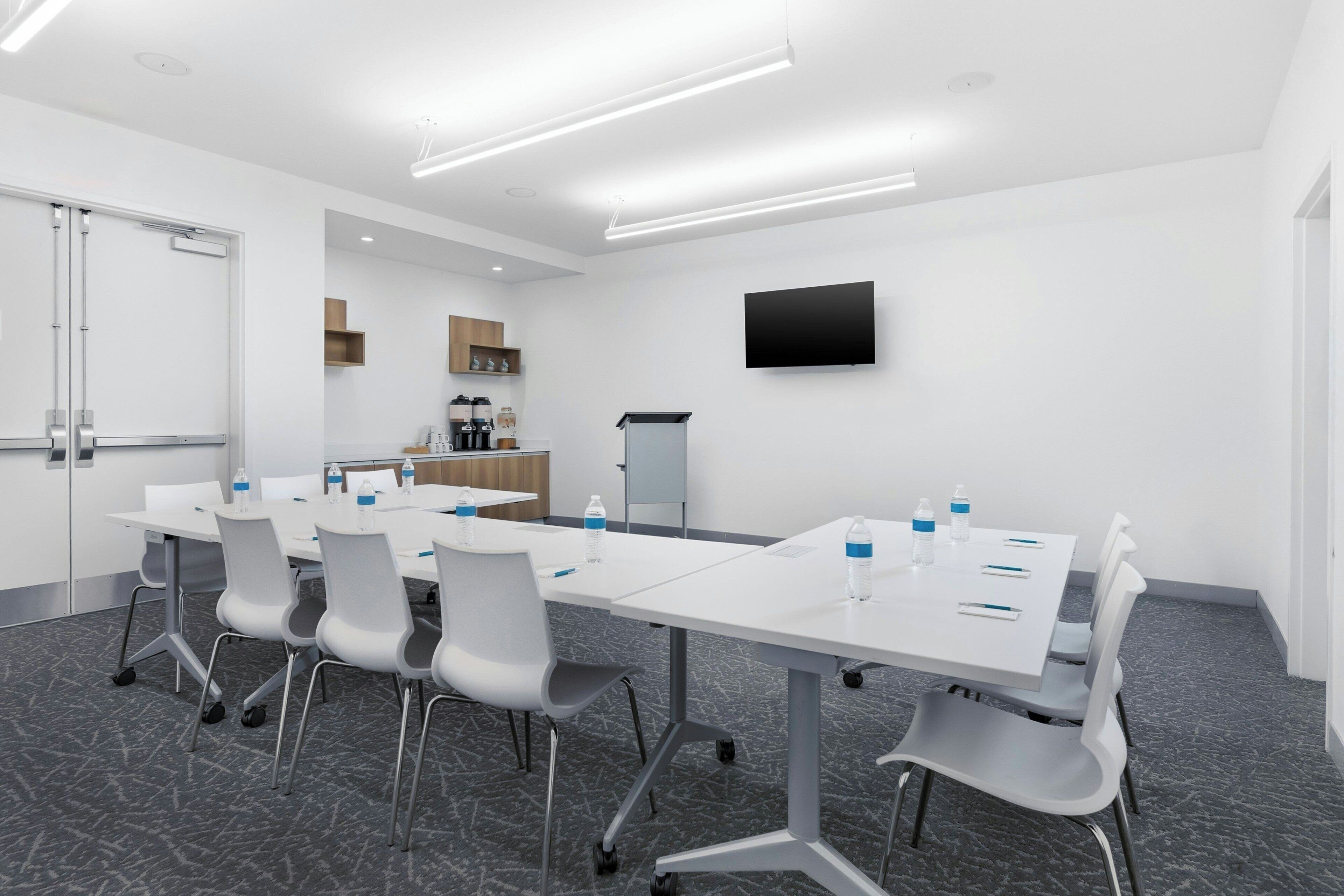 A conference room with a long table, chairs, a flat-screen TV, and a kitchenette area.