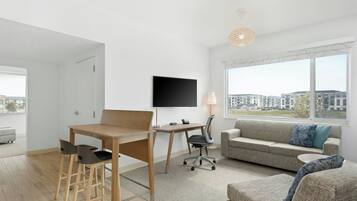 Un moderno salón con una mesa de madera, dos sillas, un sofá gris, un televisor y un ventanal con vistas a edificios.
