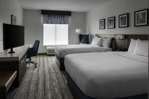 A hotel room with two beds, a desk with a TV, a chair, a lamp, and framed pictures on the wall.