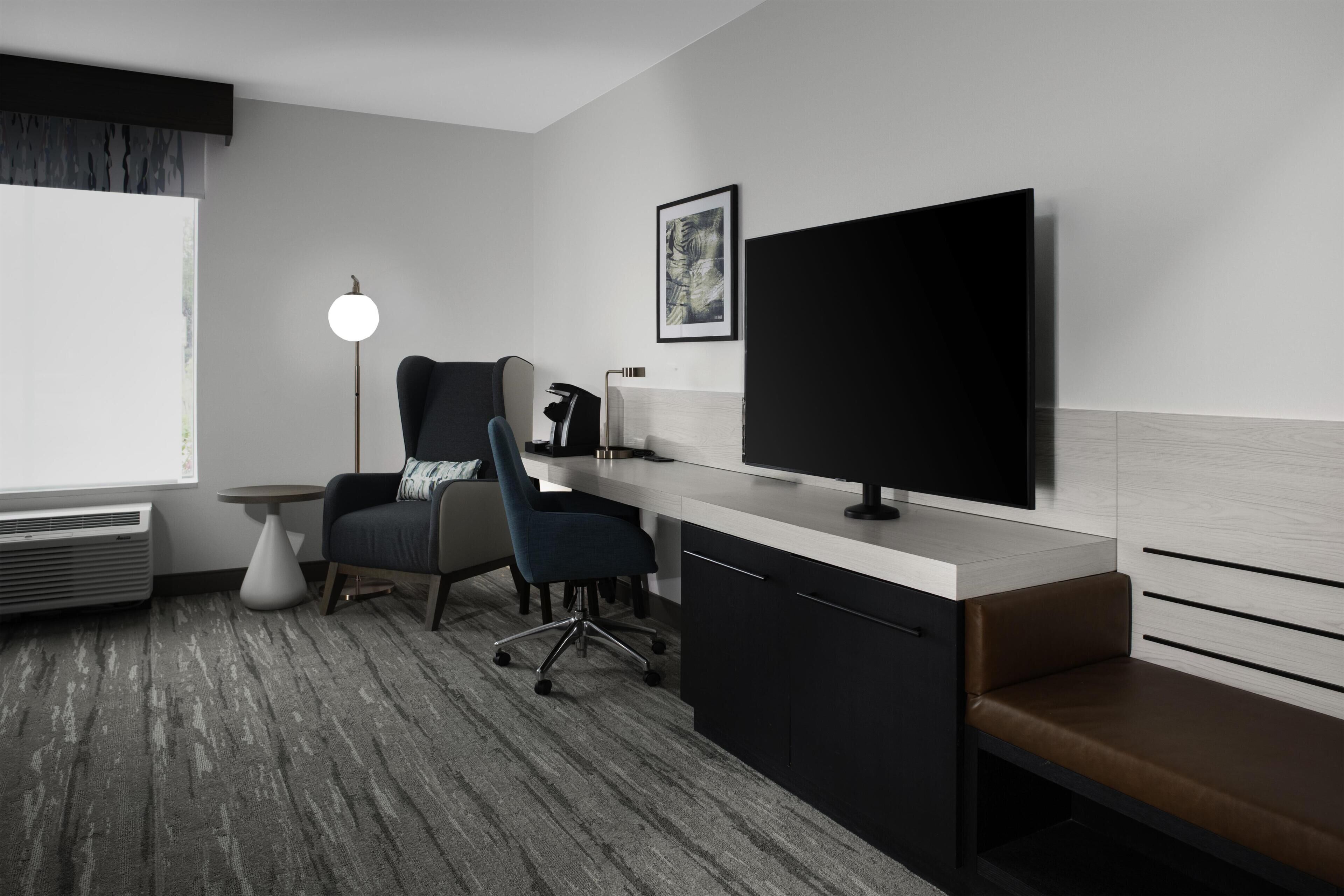 A modern hotel room with a large flat-screen TV, a desk with a chair, a small round table, a bench, and a floor lamp.