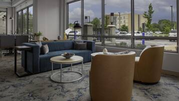 A modern hotel lobby with a blue sofa, round coffee table, and two armchairs. Large windows offer a view of the parking lot and buildings outside.