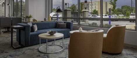 A modern hotel lobby with a blue sofa, round coffee table, and two armchairs. Large windows offer a view of the parking lot and buildings outside.
