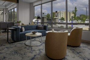 A modern hotel lobby with a blue sofa, round coffee table, and two armchairs. Large windows offer a view of the parking lot and buildings outside. - Hilton Garden Inn Jackson (Jackson)
