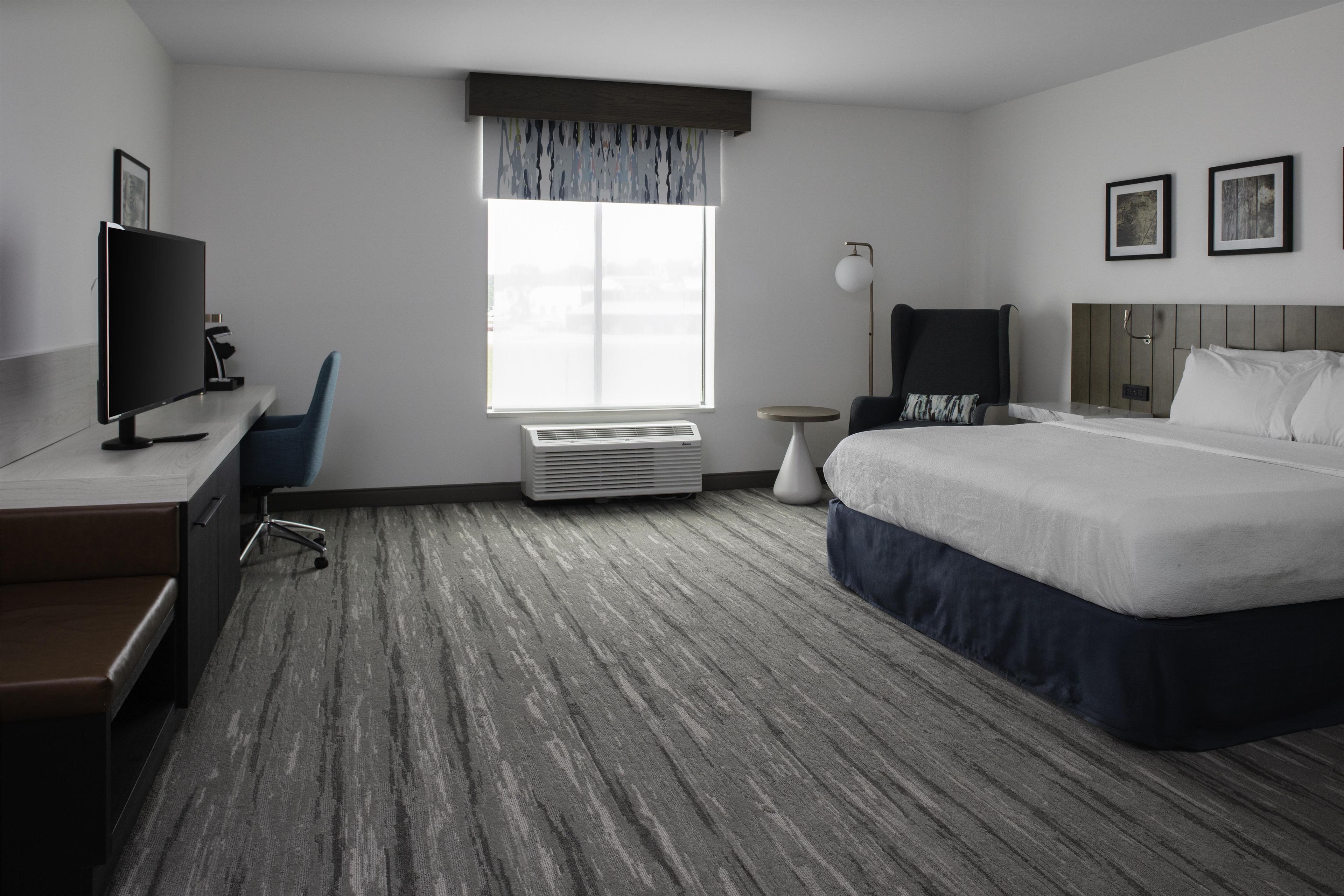 A hotel room with a bed, desk, chair, TV, air conditioning unit, and a window with curtains.