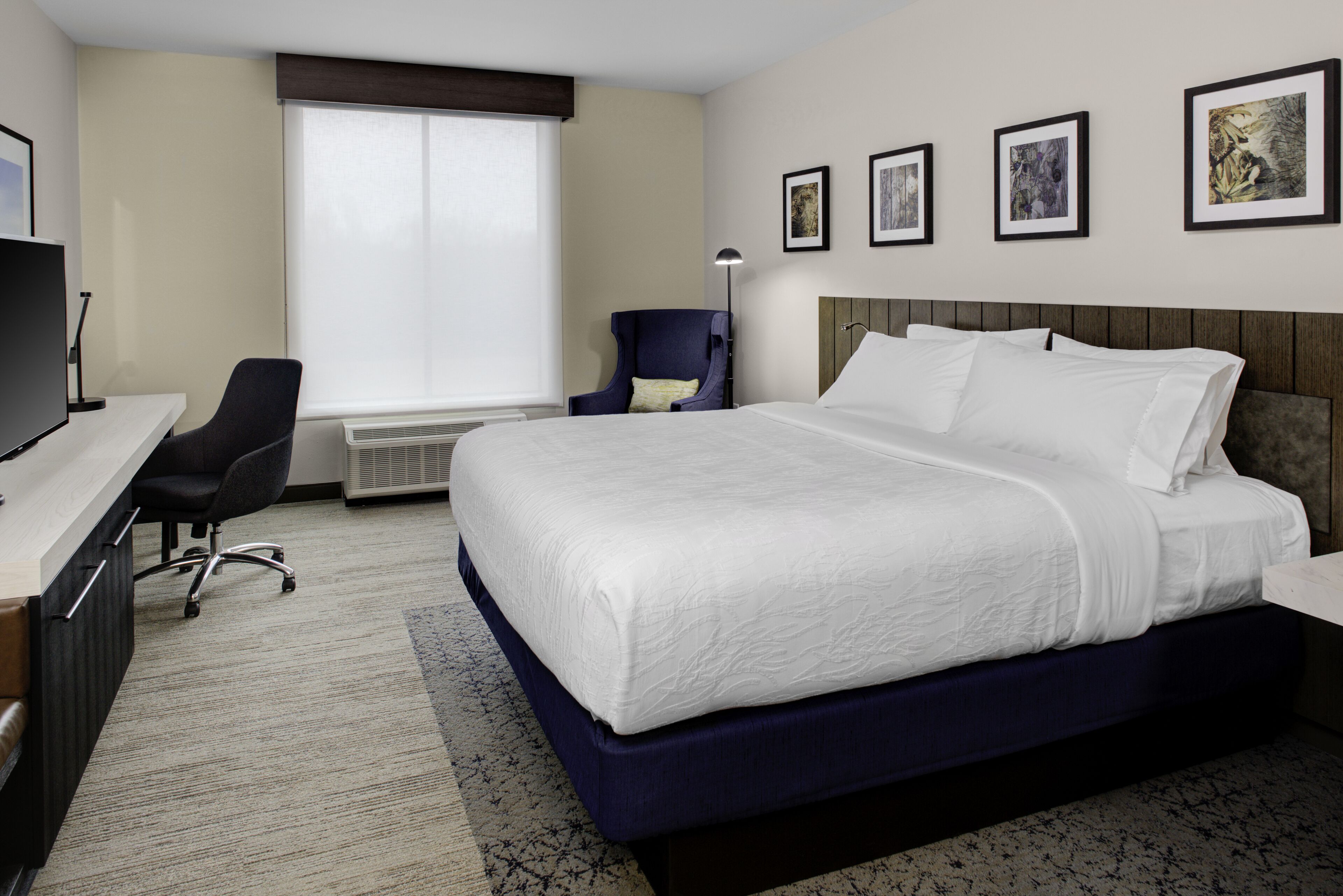 A hotel room with a large bed, a desk with a computer, a chair, and a window with blinds.
