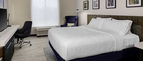 A hotel room with a large bed, a desk with a computer, a chair, and a window with blinds.