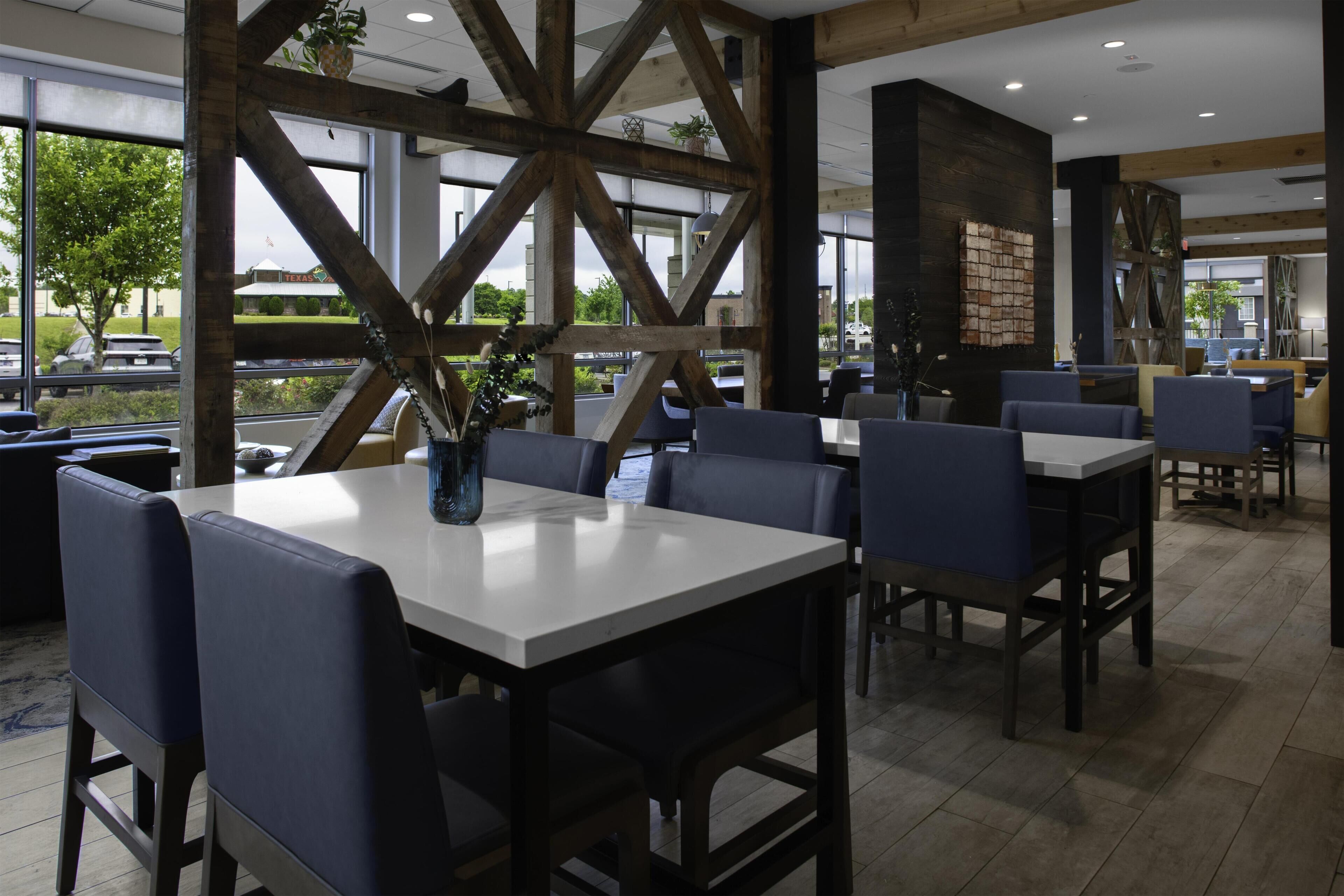 Een restaurant met een modern interieur, met zichtbare houten balken, grote ramen en diverse zithoeken, waaronder tafels en hoekbanken.