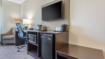 A hotel room with a flat-screen TV mounted on the wall, a desk with a coffee maker, a mini-fridge, and a chair.