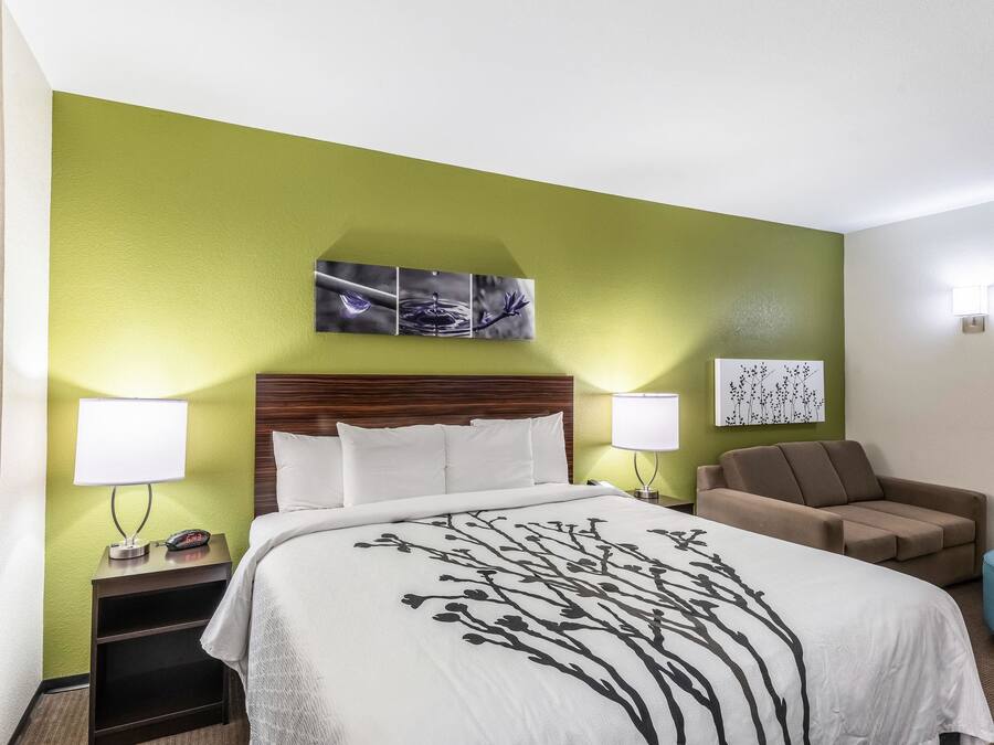 A hotel room with a bed, two bedside lamps, a nightstand, a sofa, and a wall art piece above the bed.