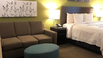 Ein Hotelzimmer mit Sofa, Hocker, einem Bett mit weißer Bettwäsche, einem Nachttisch mit Lampe und einem Wandbild.
