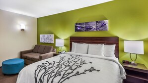 A hotel room with a bed, a blue ottoman, a brown armchair, a nightstand with a lamp, and a wall-mounted artwork. - Sleep Inn & Suites Fort Worth - Fossil Creek (Fort Worth)