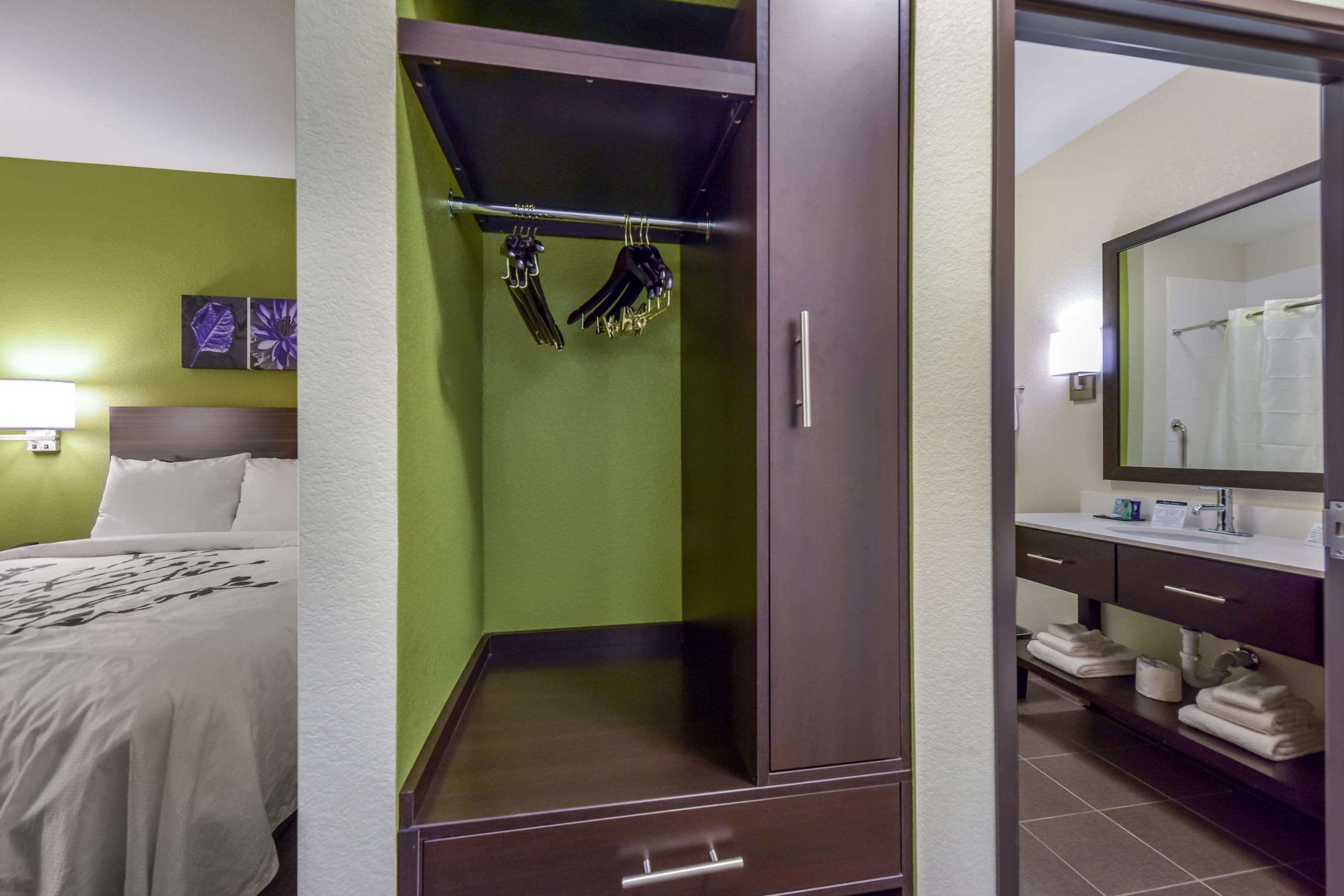 A hotel room with a bed, a wardrobe with hanging clothes, a bathroom with a sink and mirror, and a green wall.
