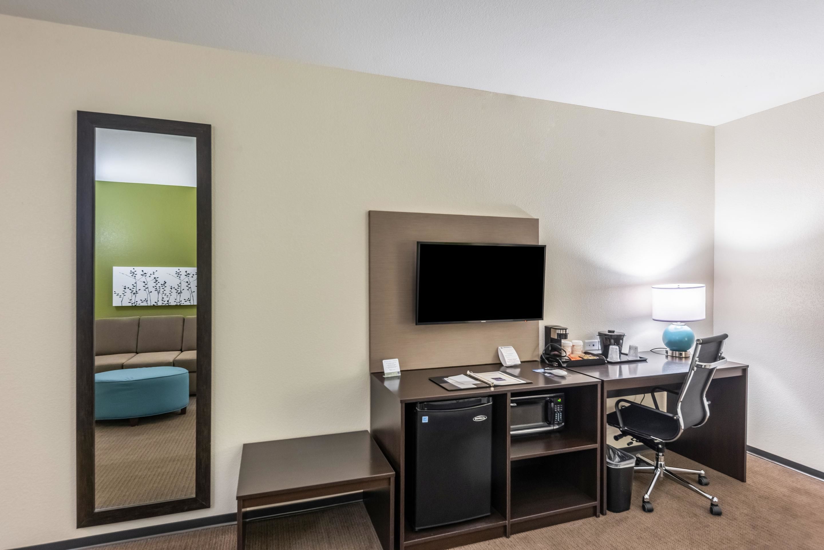 A hotel room with a desk, a flat-screen TV, a small refrigerator, a trash bin, a lamp, a chair, and a large mirror.