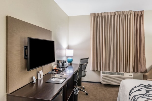 A hotel room with a flat-screen TV, a desk with a lamp, a chair, an air conditioner, and a bed.