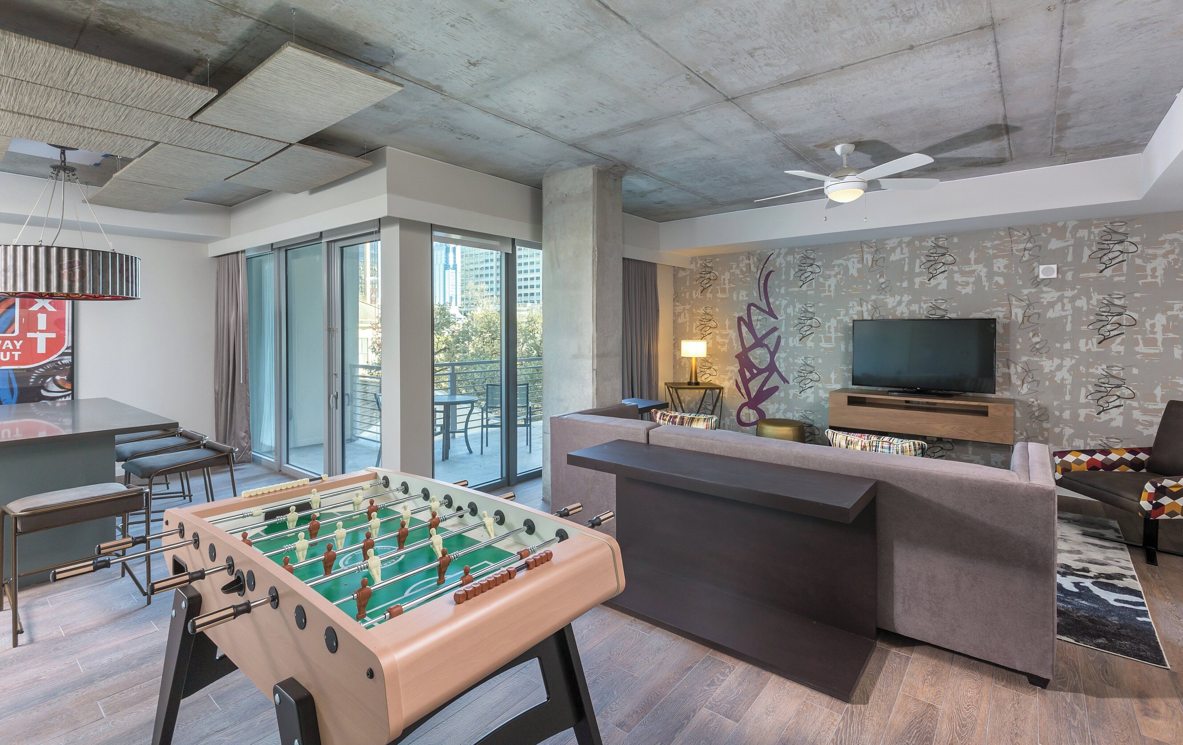 A modern living room with a foosball table, a flat-screen TV, and a balcony with outdoor seating.