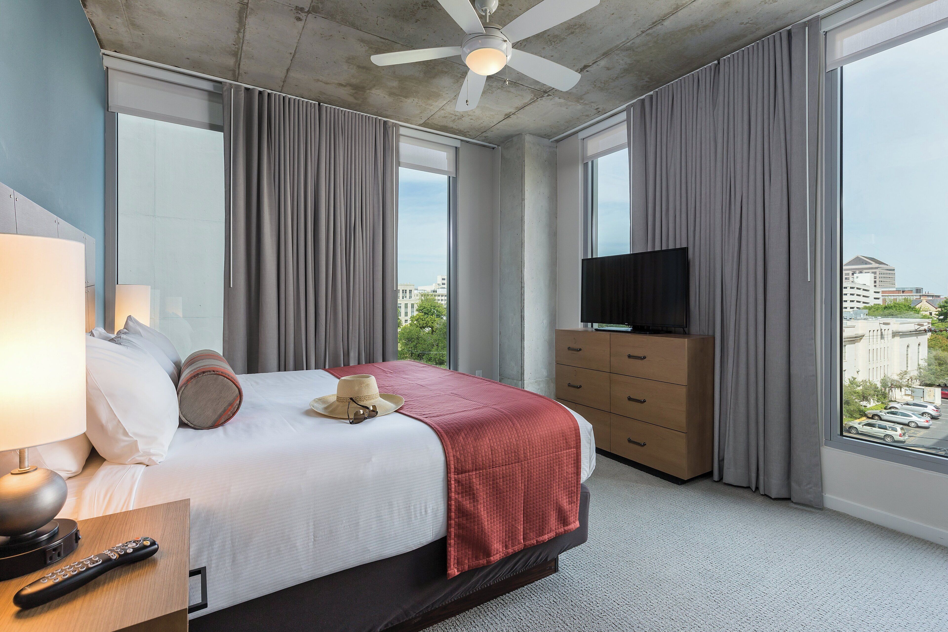 Une chambre d’hôtel avec un grand lit, une télévision, une commode et une vue sur la ville grâce à de grandes fenêtres.