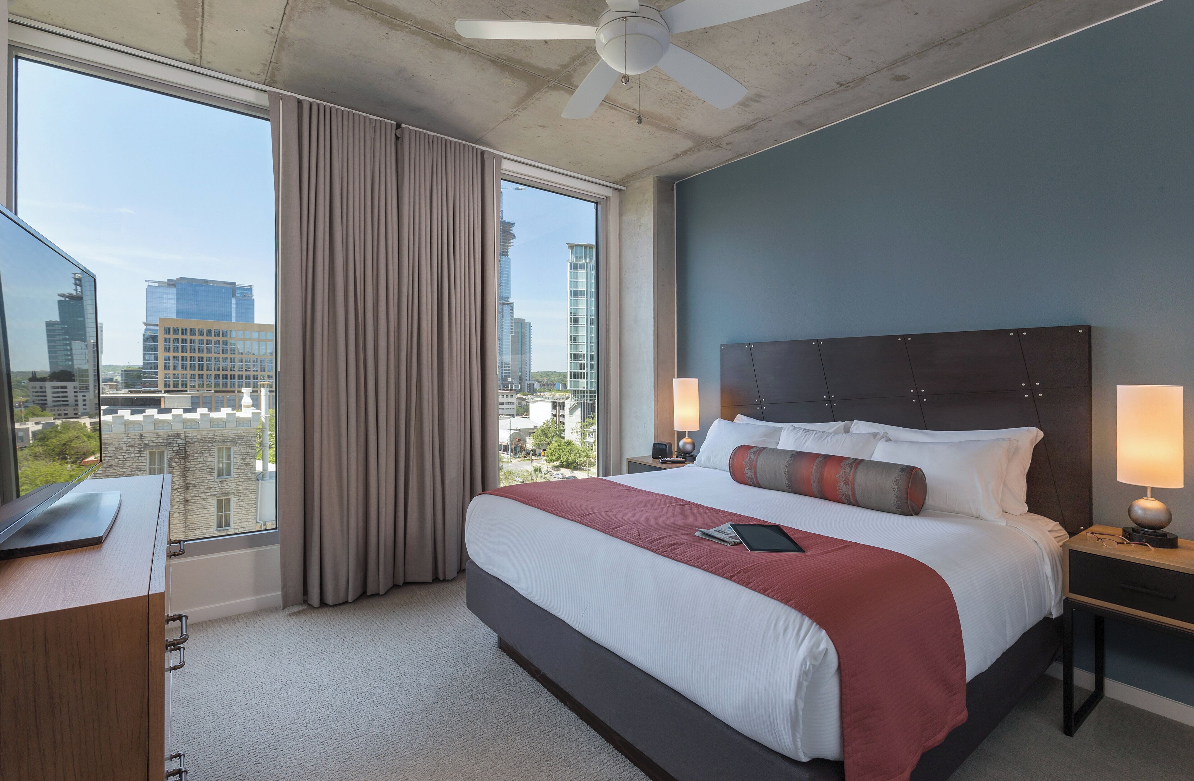 A hotel room with a large bed, a desk, a chair, a nightstand, a lamp, and a view of the city through large windows.