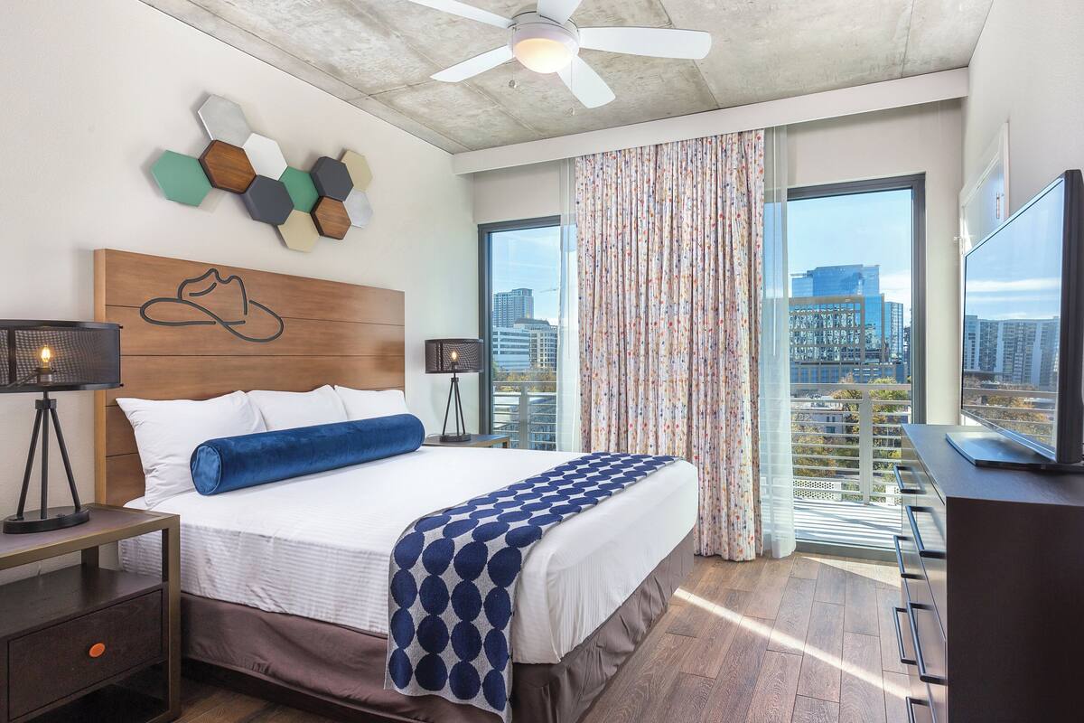 A modern hotel room with a large bed, a ceiling fan, a city view, and a desk with a TV.