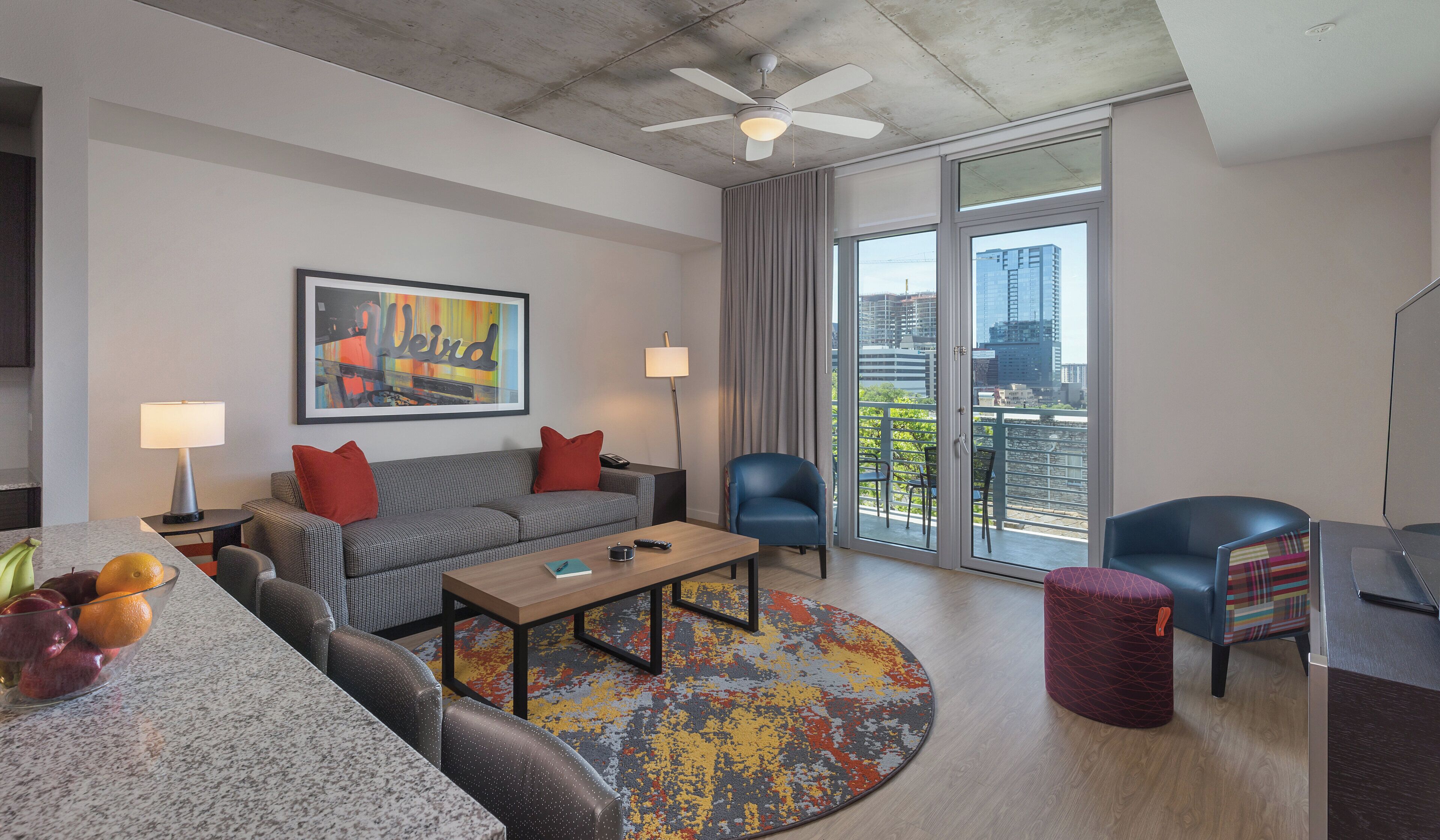 Een moderne hotelkamer met een grijze bank, rode kussens, een salontafel, een blauwe fauteuil, een rood voetenbankje, een kleurrijk vloerkleed en een groot raam met uitzicht op de stad.