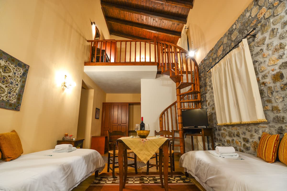 A hotel room with two beds, a dining table, a wooden staircase, and a stone wall.