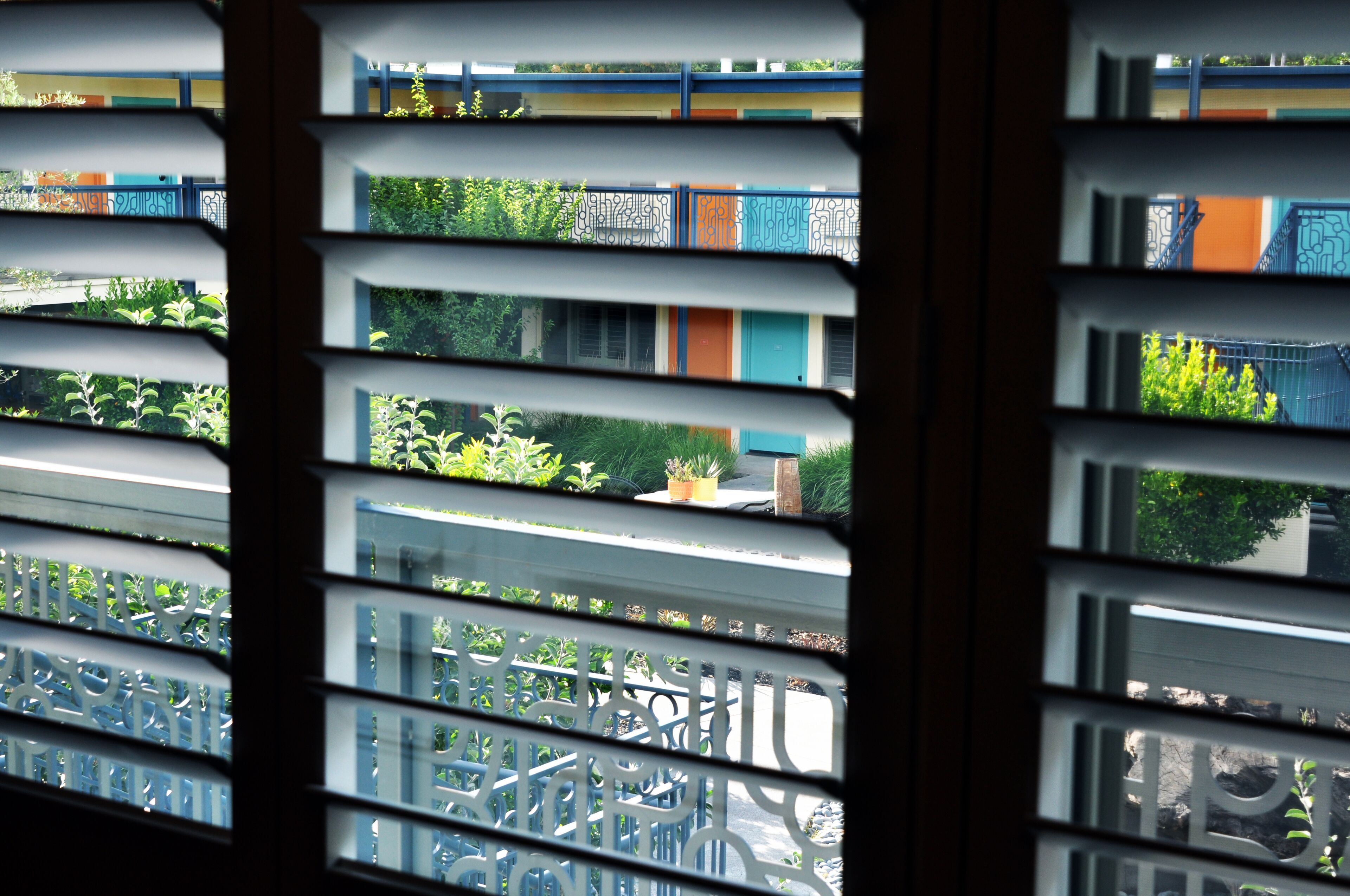Une vue à travers une fenêtre ornée d’une grille métallique, dévoilant un balcon avec des plantes et un immeuble aux portes colorées.