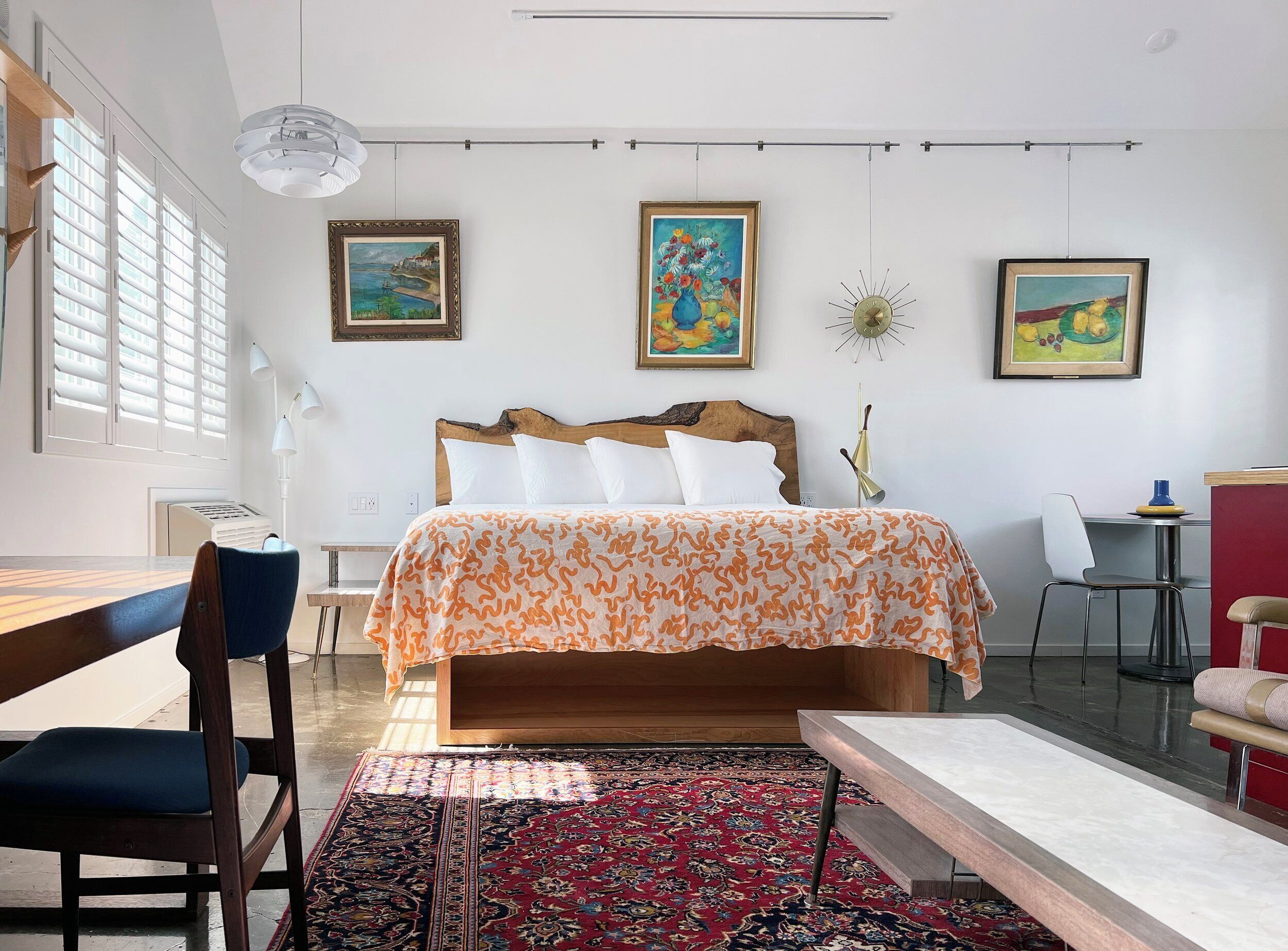 Un dormitorio moderno con una cama grande, un escritorio y una silla. Hay tres cuadros en la pared y un ventanal con persianas.