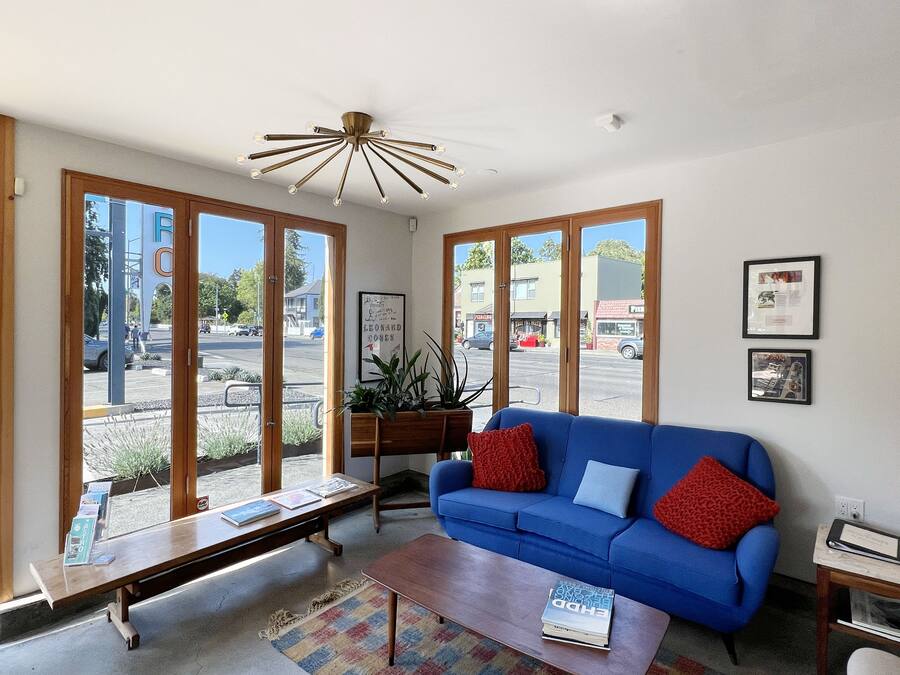 A living room with a blue sofa, a wooden coffee table, and a bench. There are large windows, a ceiling fan, and a plant on a wooden stand.