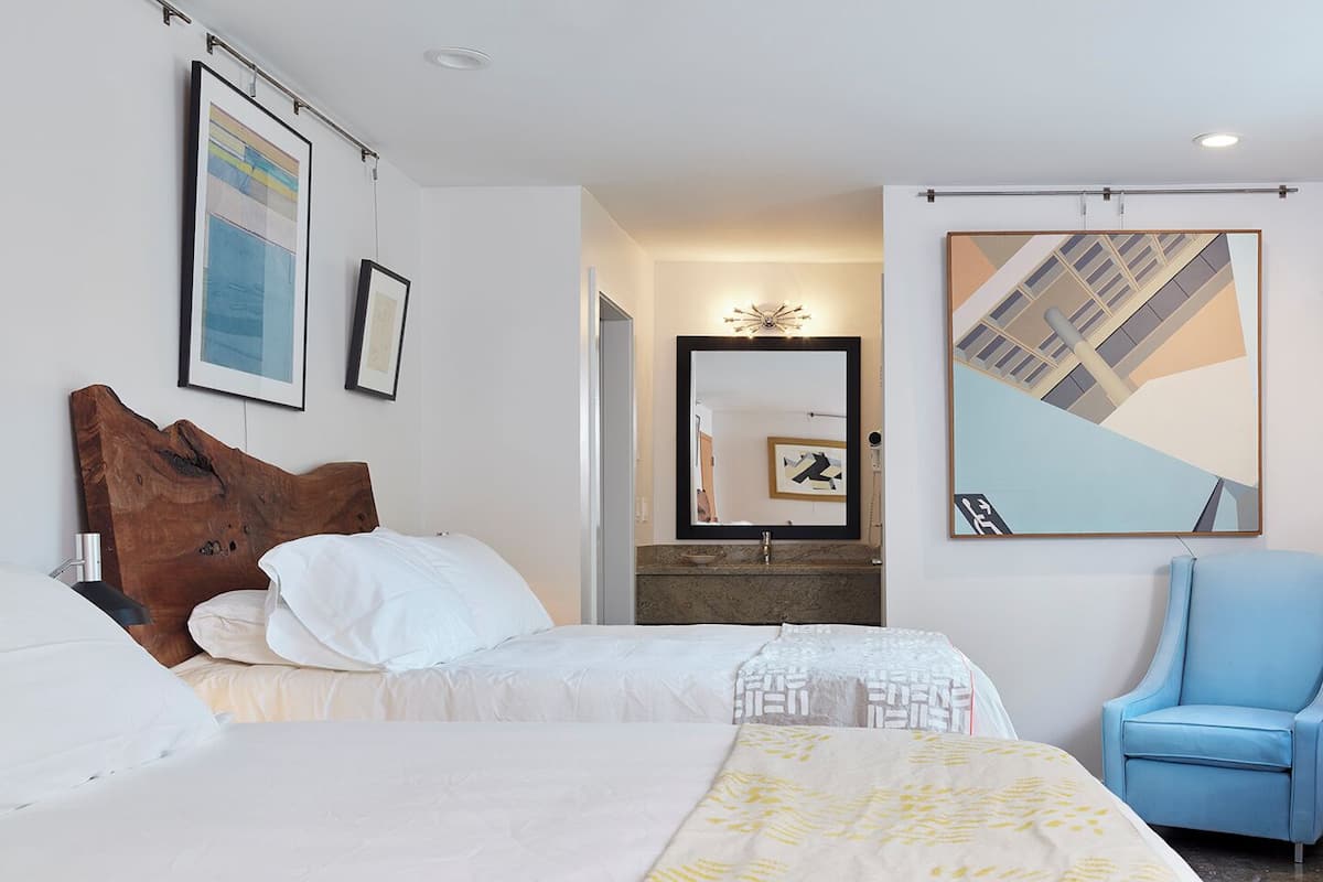 Habitación de hotel con dos camas, un escritorio, una silla, un espejo grande y obras de arte en las paredes.