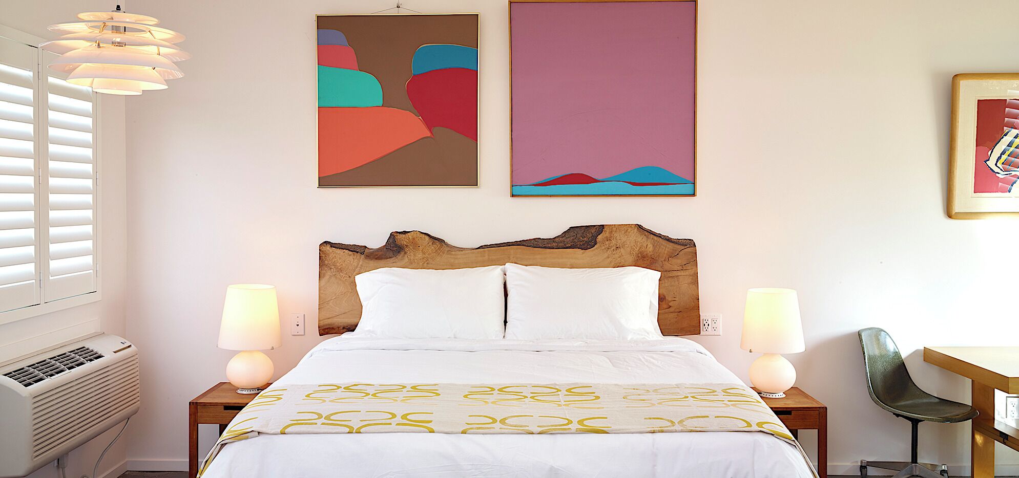 Quarto de hotel com cabeceira de madeira, cama com roupa de cama branca e padrão amarelo, duas luminárias de mesa, escrivaninha com cadeira e arte abstrata nas paredes.