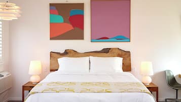 Quarto de hotel com cabeceira de madeira, cama com roupa de cama branca e padrão amarelo, duas luminárias de mesa, escrivaninha com cadeira e arte abstrata nas paredes.