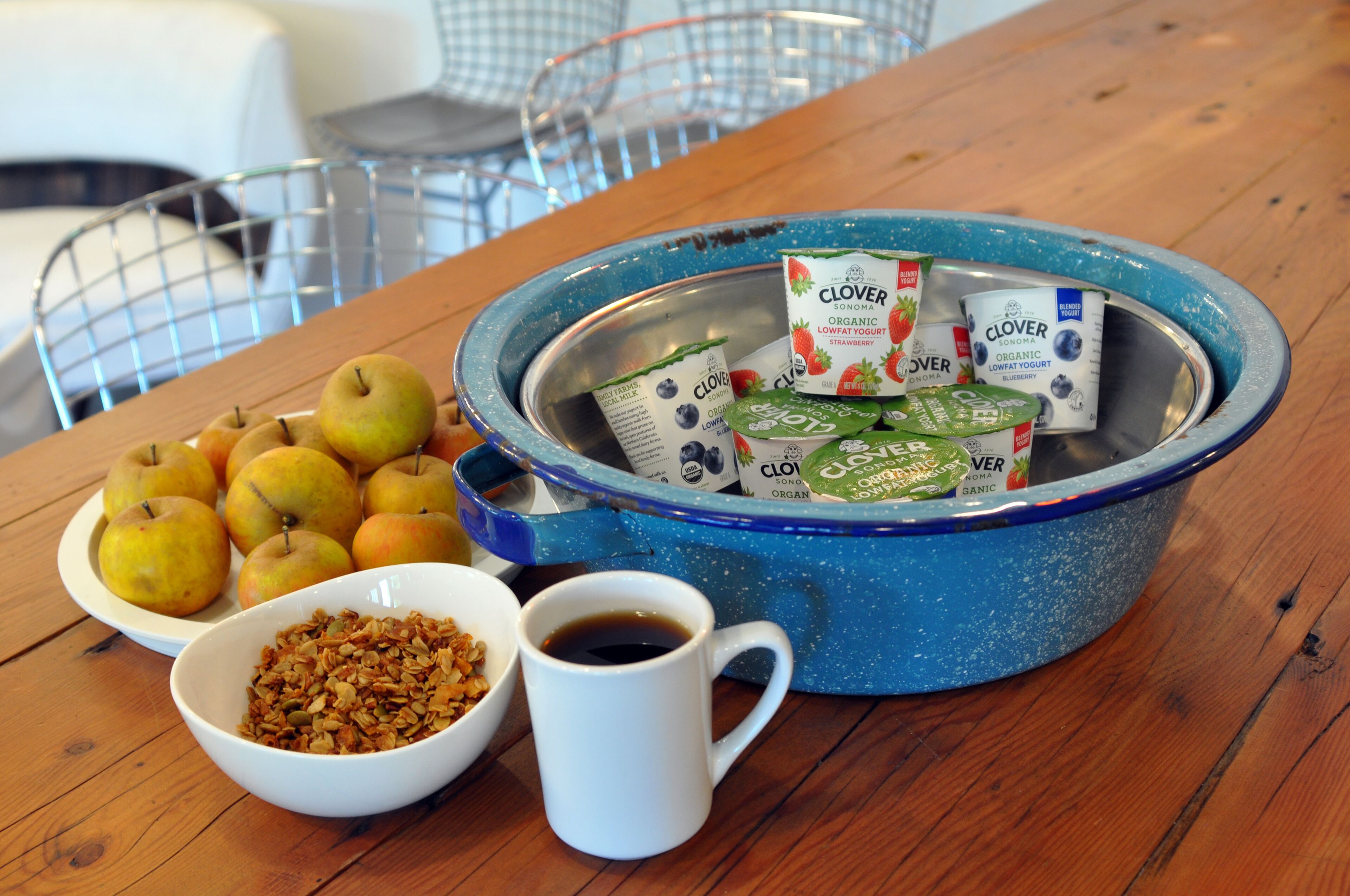 Una mesa de desayuno con manzanas en un plato, granola en un bol, una taza de café y un bol azul con yogur bajo en grasa orgánico y un cartón de leche.