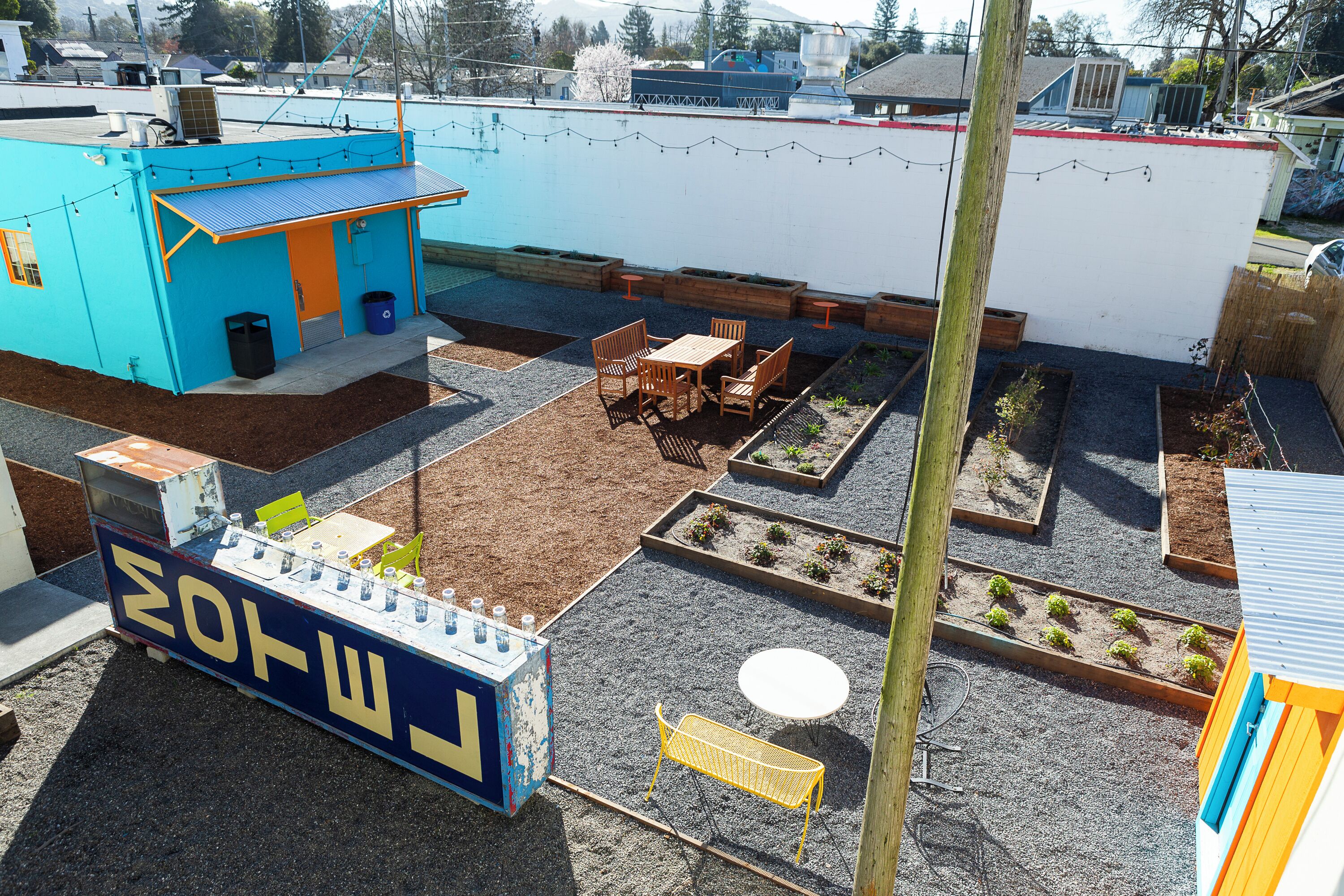 A rooftop patio with a blue building, wooden seating, raised garden beds, and a small table.