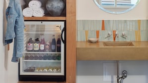 A bathroom with a wooden vanity, a round mirror, a built-in sink, and a glass-fronted refrigerator. - The Astro (Santa Rosa)