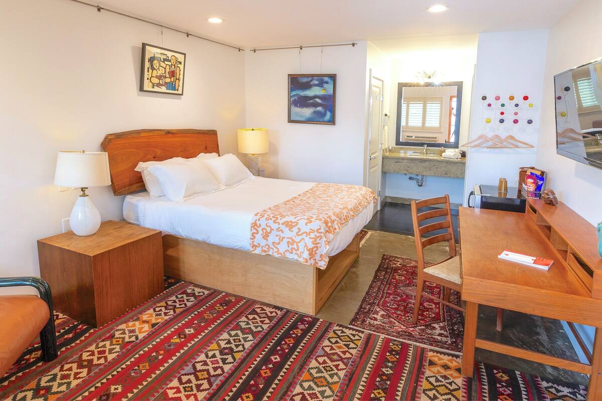 Habitación de hotel con cama, mesita de noche, lámpara, silla, escritorio y un área de baño pequeña.