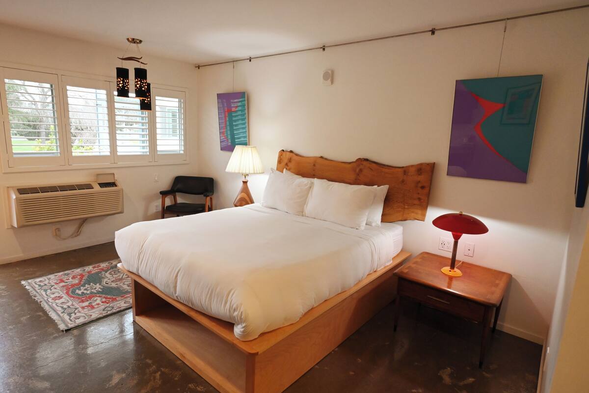Un dormitorio con cama de madera, ropa de cama blanca, una mesita de noche con lámpara, un radiador y una ventana con persianas.